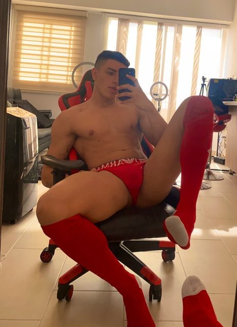 A muscular young man poses for a mirror selfie in a gaming chair wearing red underwear and knee-high red football socks.