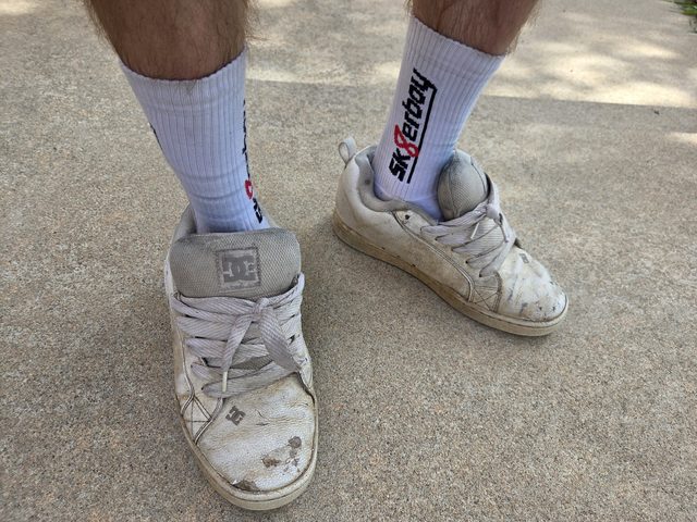 A person stands outdoors wearing white "SK8erboy" socks and dirty white sneakers.