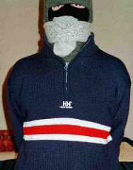 A person wears a blue quarter-zip sweater, a grey balaclava, and a green beanie indoors.