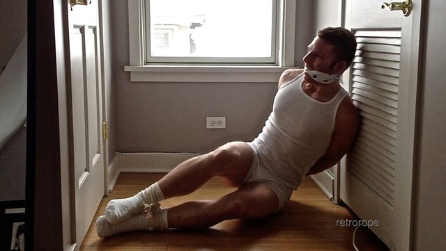A muscular man in a tank top and briefs is tied with rope and gagged, leaning against a door indoors.