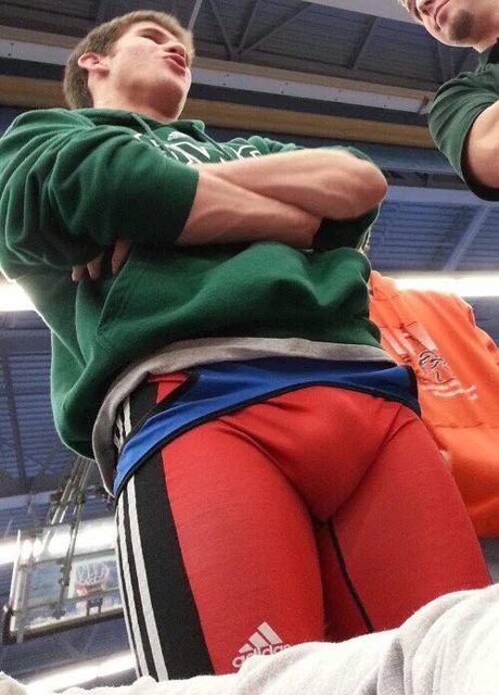 A muscular man in a green hoodie and red athletic leggings stands in a gym.