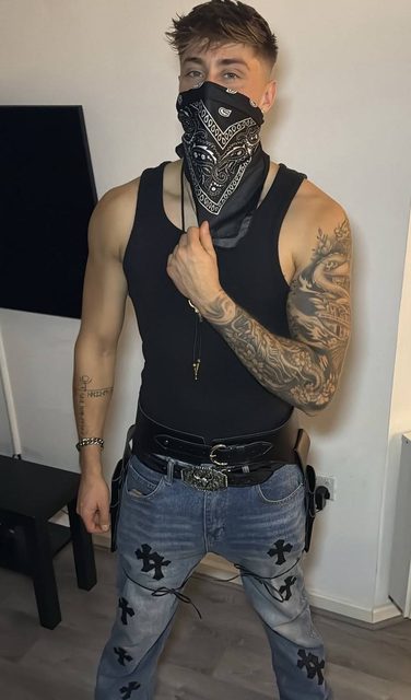 A muscular, tattooed man wearing a black tank top and jeans posed with a heavy leather utility belt and a face-obscuring bandana.