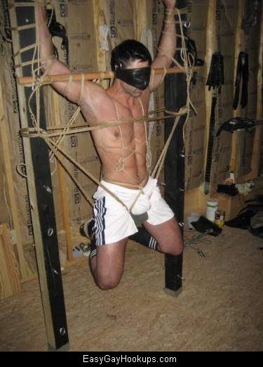 A muscular man is suspended in rope bondage, wearing a leather mask and sportswear.