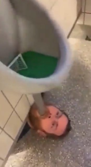 A man's face is visible on the floor next to a urinal in a public restroom.