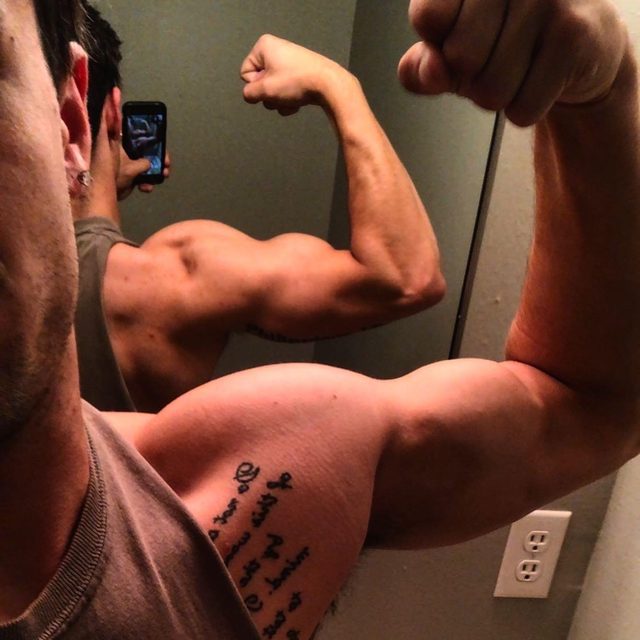 A muscular man with tattoos flexes his bicep while taking a mirror selfie.