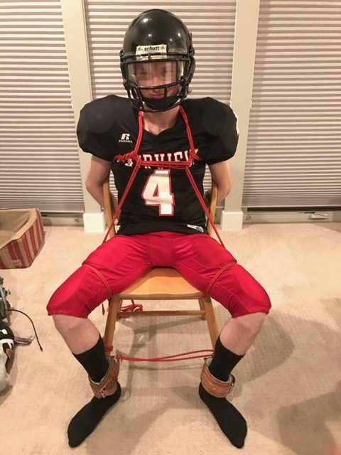 A young man in a full American football kit is bound to a wooden chair with red ropes and leather ankle cuffs.
