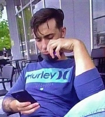 A man with a beard and tattoos sits outdoors looking at his phone.