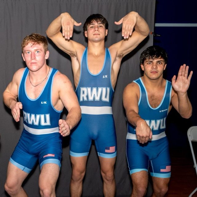 Three young men in blue wrestling singlets pose together in an indoor setting.