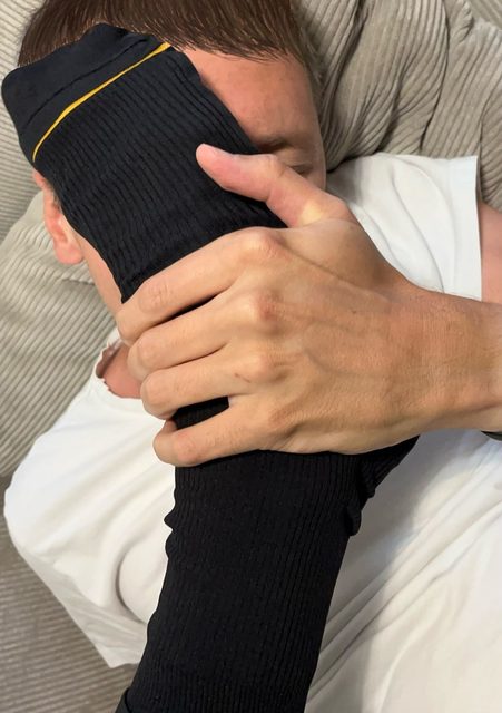 A man lies back while a foot in a black ribbed sock is pressed firmly against his face and held in place by a hand.