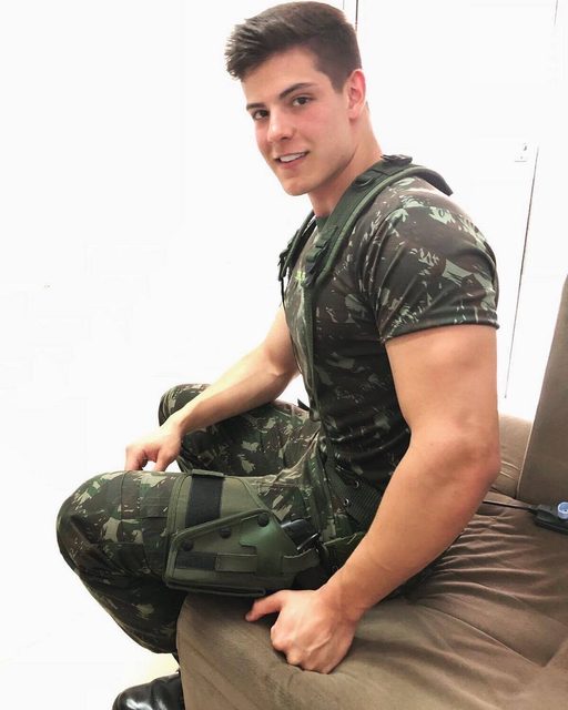 A muscular man in a military uniform and tactical gear sits on a couch and smiles.