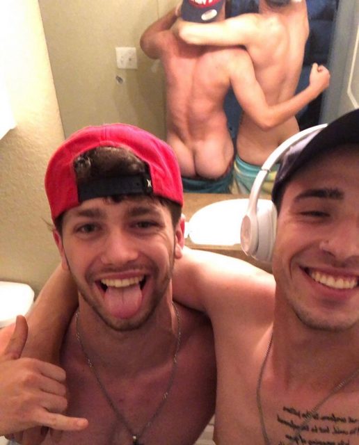 Two athletic young men pose for a shirtless bathroom selfie while their reflection reveals one showing his bare buttocks.