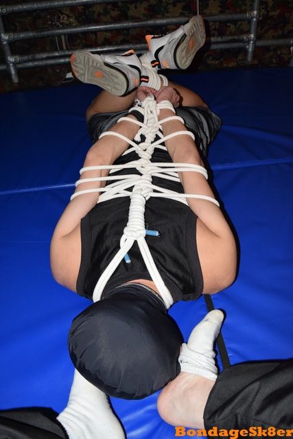 A man is suspended upside down, bound with white rope, wearing a black hood and sneakers.