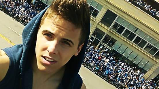 A young man takes a selfie outdoors in front of a large crowd.