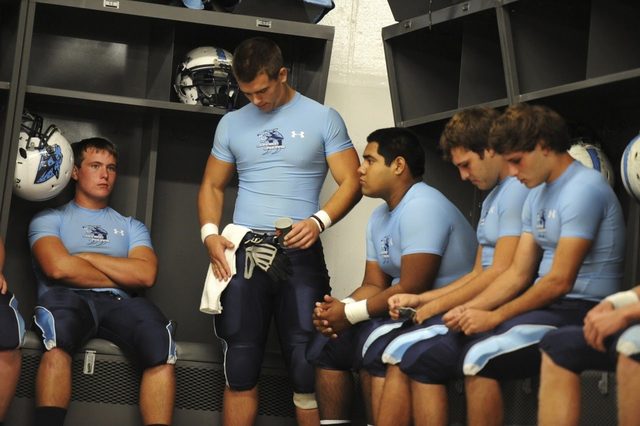 A group of athletic young football players wearing sky blue compression tops and padded football pants inside a locker room.