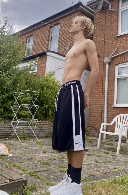 A lean, shirtless young man with blonde hair poses in a backyard wearing basketball shorts, black socks, and white sneakers.