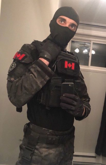 A man poses for a selfie wearing a full tactical uniform with a black balaclava and camouflage gear.