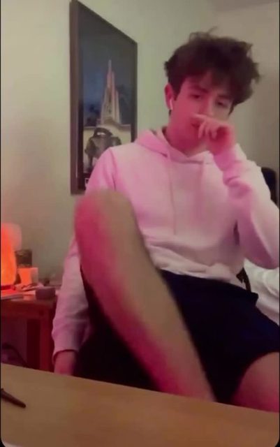 A young man in a pink hoodie and dark shorts sits casually in a room.