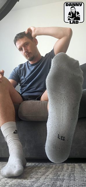 A man in a t-shirt and gym shorts sits on a sofa, extending a white crew sock-covered foot towards the camera in a POV perspective.
