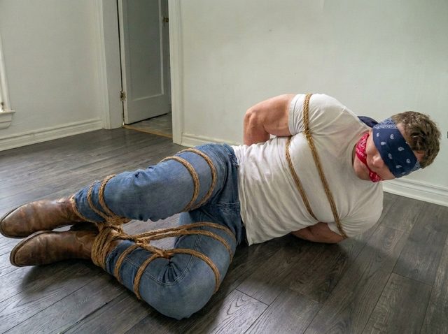 A muscular man in denim and a white t-shirt lies on a wooden floor, bound with thick ropes, blindfolded, and gagged.