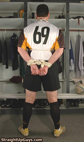 A man in a rugby uniform stands bound with rope behind his back in a locker room.