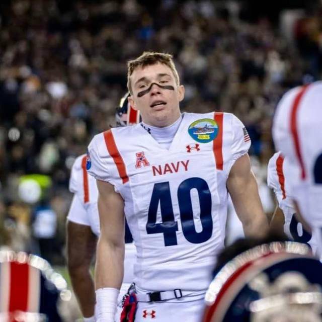 A young, muscular football player in a white Navy jersey and shoulder pads stands on the field during a game.