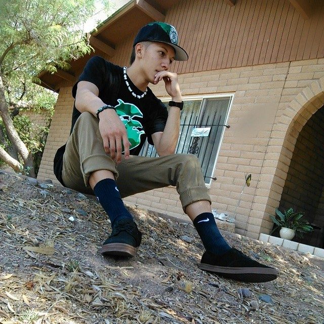 A young man sits outdoors on a dirt slope, wearing a cap, t-shirt, and sneakers.