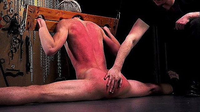 A naked submissive man in a full split is restrained in a wooden pillory while a dominant touches his heavily reddened back and buttocks in a dungeon.
