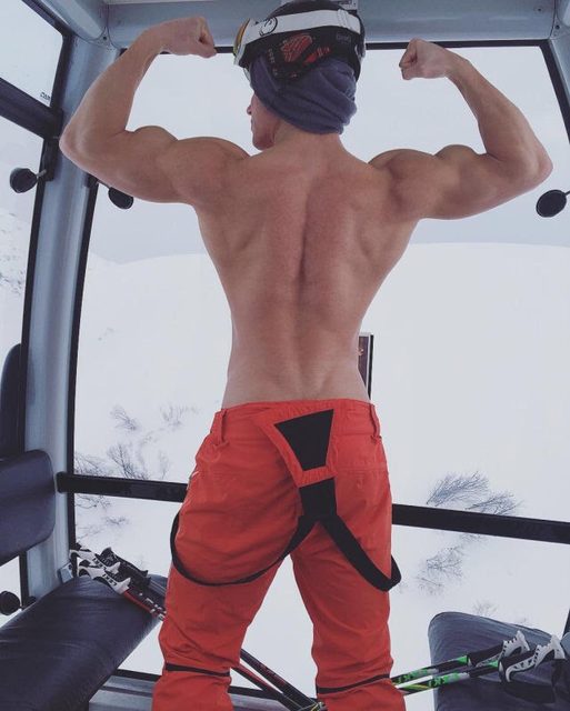 A muscular man in ski pants and a helmet flexes his back and arms in a gondola with a snowy view.