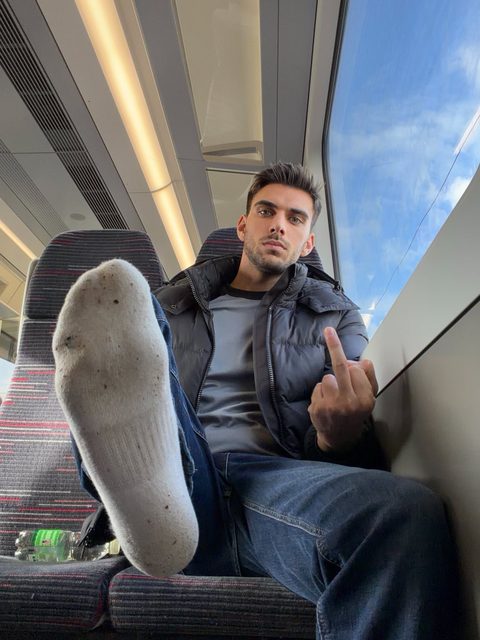 A bearded man sitting on a train holds up his middle finger while extending a dirty white sock toward the camera.