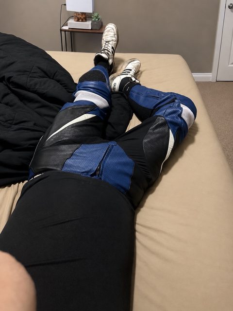 A person wearing blue and black motorcycle leathers and white sneakers lies on a bed.