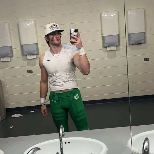 A muscular man wearing sportswear and a cap takes a mirror selfie in a public bathroom.