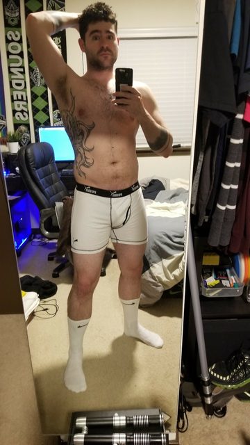 A man in boxer briefs and socks takes a mirror selfie in his bedroom.