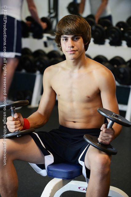 A shirtless young man with a smooth chest sits on a bench in a gym holding dumbbells.