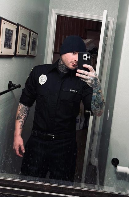 A man wearing a black uniform shirt, cap, and tattoos takes a mirror selfie indoors.