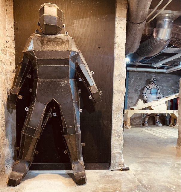 A large metal bondage structure stands in the foreground, with another masked figure and wooden BDSM gear visible in an industrial basement setting.