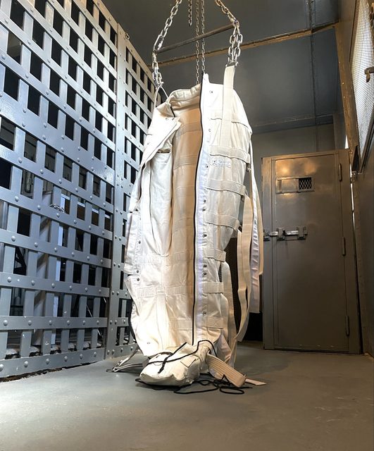 A straightjacket hangs from chains in what appears to be a prison cell.