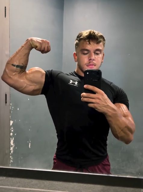 A muscular man takes a mirror selfie while flexing his bicep in a gym.