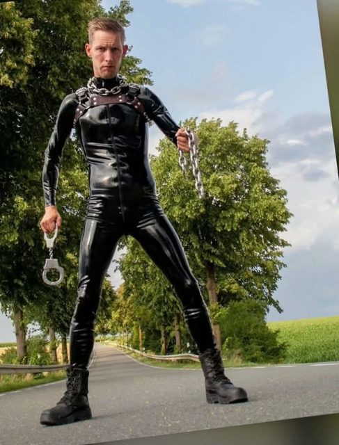 A man in a black latex suit, leather harness, and boots poses with chains and handcuffs on an outdoor road.