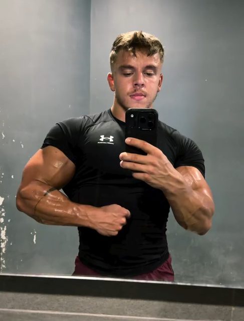 A muscular man in sportswear takes a mirror selfie in a gym or locker room.