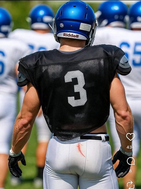 A muscular football player in a black jersey and white pants with a blue helmet stands on an outdoor field.