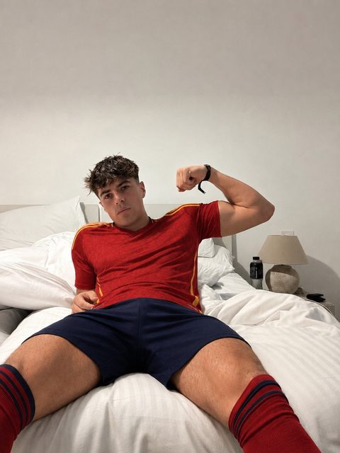 A muscular man in sportswear poses on a bed in a bedroom.