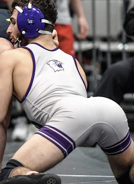 A muscular man in a wrestling singlet and headgear takes a stance on a wrestling mat.