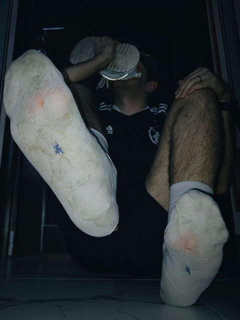 A man holds a sneaker to his face, showing his worn socks and hairy legs.