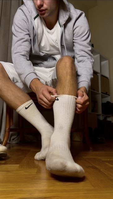 A man in a grey hoodie and white shorts adjusts his white socks.