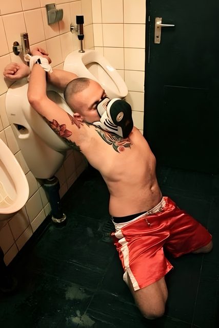 A man is restrained to a public urinal with his hands tied behind his back, wearing red athletic shorts and sneakers.