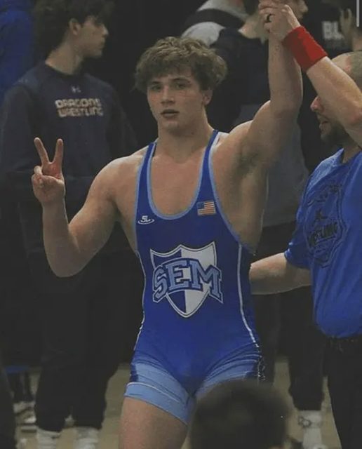 A young muscular man in a blue wrestling singlet makes a victory sign in a gym setting.
