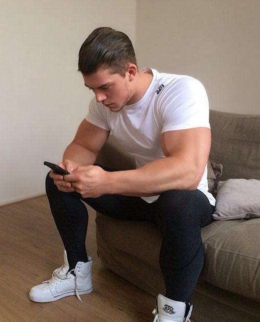 A muscular man in a white t-shirt, black leggings, and white high-top sneakers sits on a couch while looking at his phone.