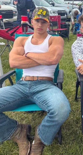 A young muscular man in a tank top, jeans, and cowboy boots sits outdoors.