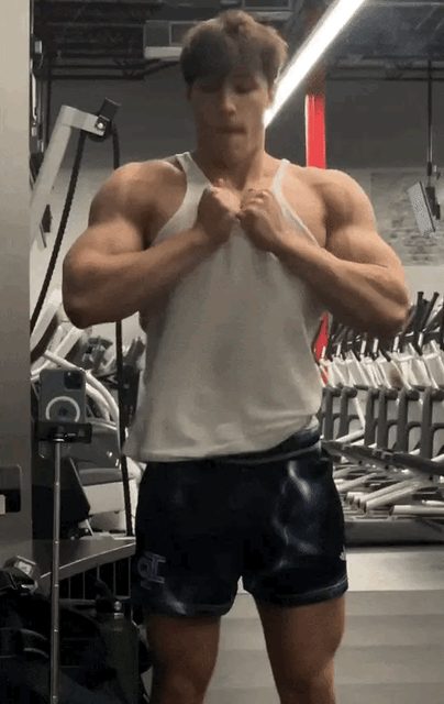 A muscular man in a tank top and shorts flexes in a gym.