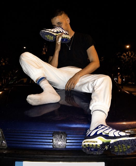 A young man sits on a car hood at night, holding a sneaker up to his face.
